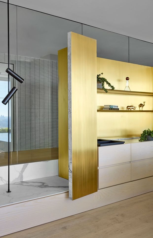 A refined contemporary interior featuring a pale timber built-in cabinetry wall with open display shelving, a dramatic brushed gold-toned partition panel, and a white marble-lined recessed area. Full-height glazing with horizontal blinds, a sculptural black floor lamp, and curated decor complete the space. The image emphasizes custom joinery, layered partitions, and warm neutral materials.