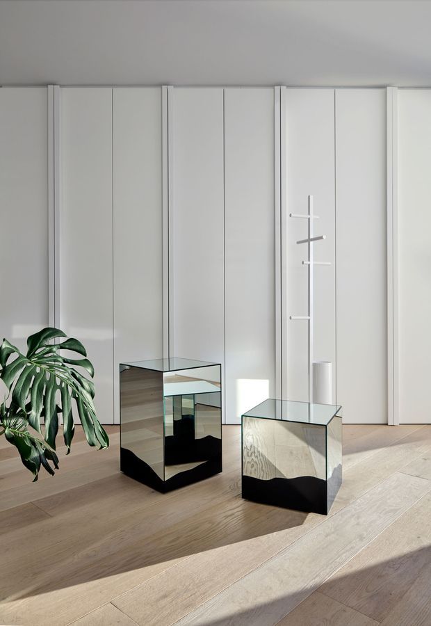 A bright minimalist interior with full-height white built-in wall panelling or cabinetry, pale timber flooring, and two transparent glass display cubes with black bases. A large leafy indoor plant sits at the left, while strong sunlight casts geometric shadows across the floor and furniture. A slim sculptural wall-mounted coat rack and simple white vase add restrained decorative elements.