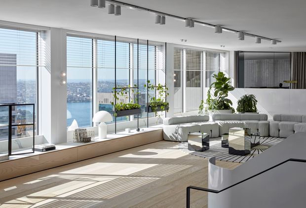 Bright, spacious contemporary living room with floor-to-ceiling glazing overlooking a waterfront cityscape. The room features white modular sofas, a pale timber built-in window seat, indoor plants, patterned rug, sculptural side tables, white cabinetry, and track lighting. Strong natural light creates crisp shadows across the pale timber floor.