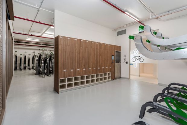 Bright, functional commercial fitness facility interior with a large timber locker and cubby storage wall, rows of exercise bikes, and distinctive overhead suspended cycling racks. The space has polished light-grey flooring, white walls and exposed services beneath a high ceiling.
