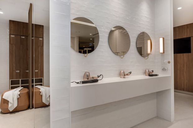 Contemporary commercial bathroom vanity area with a long white integrated counter, three sculptural basins and wall-mounted taps beneath three round mirrors. White textured wall tiles and concealed linear lighting create a clean, refined interior. A full-height mirror at left reflects adjacent timber lockers and upholstered seating, while warm timber wall panelling and a dark display screen are visible at right.