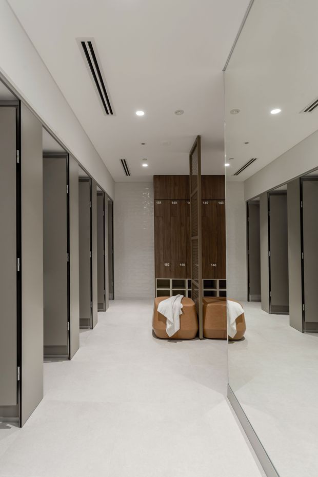 A bright, highly refined communal changing-room or dressing area with rows of grey partitioned cubicles on both sides, a full-height mirrored wall, pale seamless flooring and walls, recessed downlights, linear air-conditioning vents, and a central timber screen with built-in shelving. Two rounded tan upholstered seats with white towels sit in the centre.