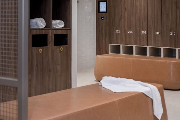 A refined locker-room or spa changing area with warm wood-grain locker cabinetry, numbered compartments, integrated open cubbies, and soft caramel-brown upholstered benches. White towels and pale tiled surfaces add contrast, while a glazed or mesh partition is visible at the left.