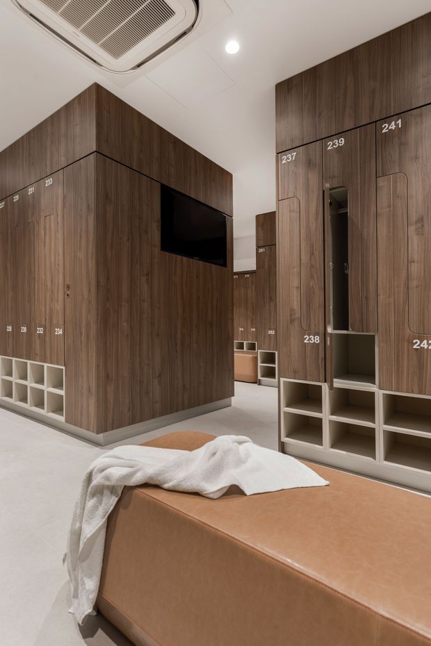 A compact, highly organised interior with floor-to-ceiling dark timber veneer storage cabinetry, integrated open cubbies, recessed handles, and numbered locker-style compartments. A wall-mounted television is integrated into the cabinetry, while a large tan upholstered bench or ottoman occupies the foreground. The space has a white ceiling with recessed lighting and a ceiling-mounted air-conditioning unit, with a light neutral floor.