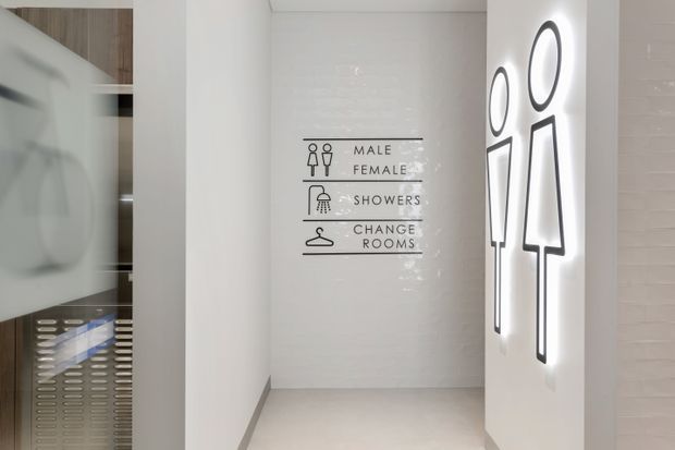 A clean, modern commercial amenities corridor with white tiled walls and pale flooring. Graphic signage identifies male and female facilities, showers, and change rooms, while large backlit male and female restroom silhouettes decorate the right wall. A partial glimpse of a shower or changing area is visible through the opening on the left.