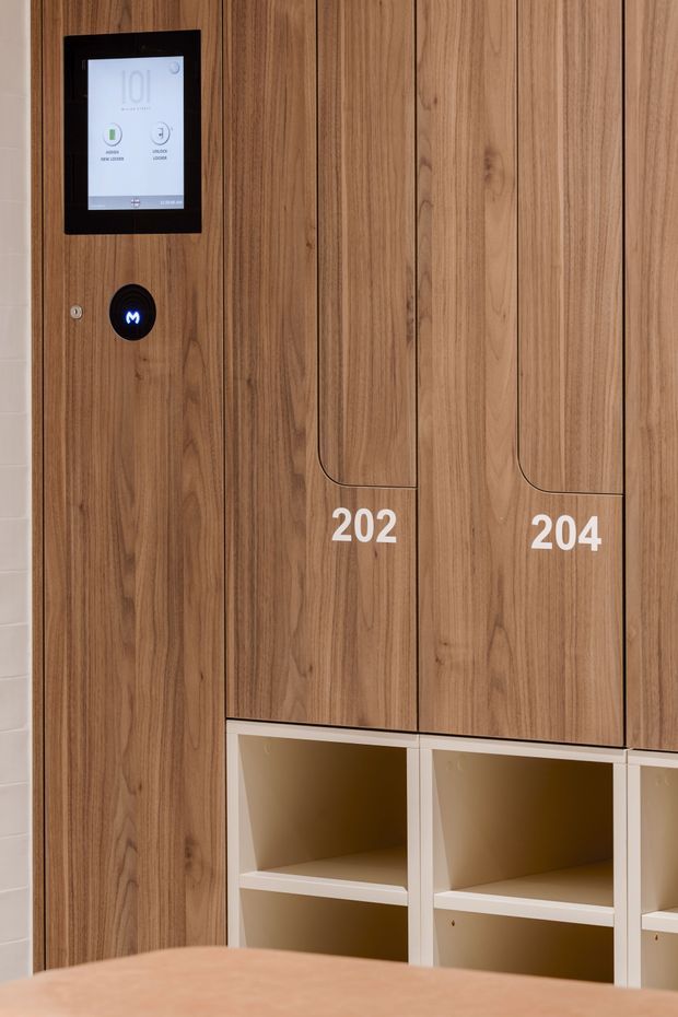 Close-up of a contemporary built-in timber locker or storage wall, with vertically grained warm wood doors labelled 202 and 204, integrated open shelving below, and a wall-mounted digital access or control panel. The cabinetry has clean recessed pulls and a refined commercial or shared-facility fit-out.