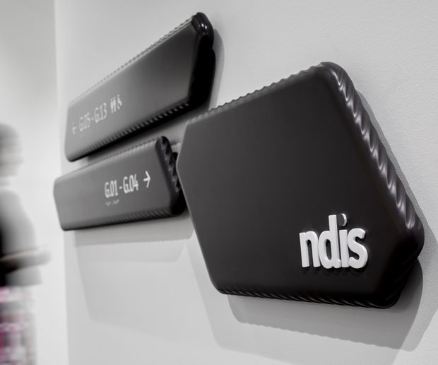 Close-up product photograph of three black padded wall-mounted or displayed protective cases, with white lettering and an “ndis” logo on the foreground case. The objects are mounted against a clean light-grey wall and lit to emphasize their sculpted edges and shadows.