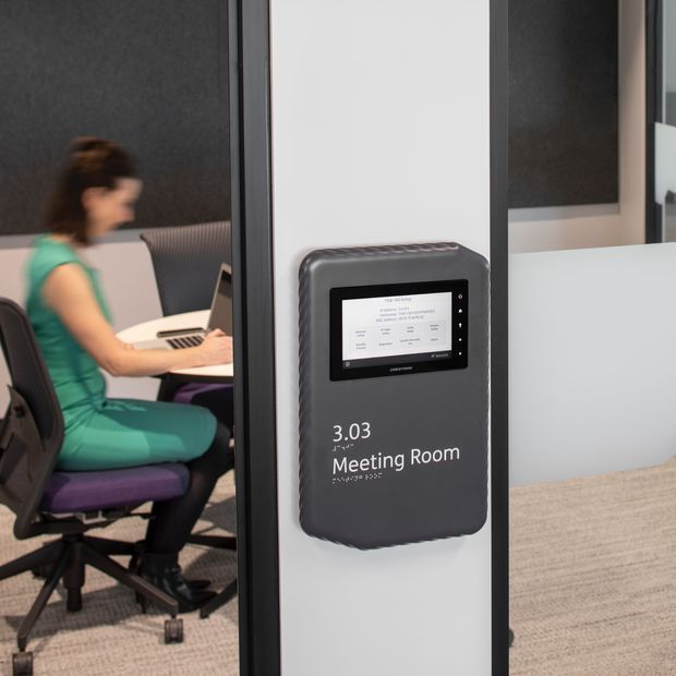 A modern office meeting-room entrance featuring a wall-mounted digital room-booking display labelled “3.03 Meeting Room.” Through the glass, a person is seated at a desk with a laptop in a contemporary workstation. The space uses dark acoustic wall panels, glass partitions with dark frames, light walls, and carpet flooring.