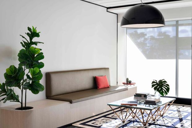 A bright, minimalist living room with a built-in upholstered bench along a pale wood joinery wall, a large black dome pendant light, expansive windows, a glass coffee table with geometric metal legs, and a patterned area rug. Indoor plants and a single red cushion provide contrasting accents.