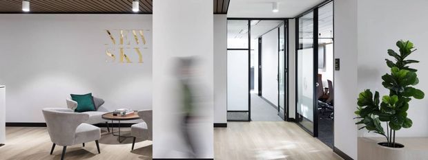 Contemporary office interior with a reception or lounge area on the left and a glazed corridor leading to private offices on the right. The space features black-framed glass partitions and doors, pale timber flooring, white walls, a dark slatted ceiling, modern lounge seating, and indoor planting.