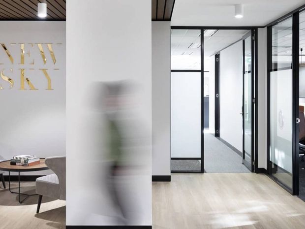 A contemporary commercial office interior with a bright reception or waiting area in the foreground and glazed, black-framed office partitions opening onto a corridor. The space features pale timber-look flooring, white walls, black skirting and framing, a dark slatted ceiling section, and minimalist furnishings. A blurred person is present, and gold wall lettering is partially visible.