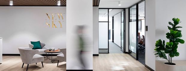 Contemporary commercial office interior with a light timber floor, white walls, black-framed glazed meeting-room partitions, and a small reception or lounge area with upholstered armchairs and a round coffee table. A feature timber-slat ceiling and indoor greenery add warmth to the otherwise clean, minimalist workspace.
