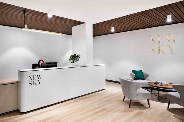 A contemporary reception lobby with a large white curved-front reception desk, wood-slat ceiling panels, light timber flooring, white walls, pendant and recessed lighting, and a small seating area with upholstered lounge chairs and a round coffee table. Gold wall lettering provides a branded feature.