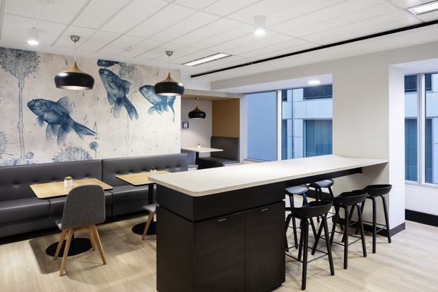 A contemporary commercial break-out or staff lounge with a large black island counter, integrated seating, bar stools, booth seating and small tables. The room features a pale timber-look floor, white walls, a suspended grid ceiling with recessed lighting, pendant lights, extensive windows, and a large monochrome fish mural.