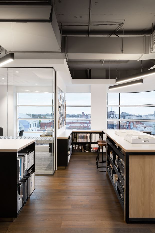 A bright contemporary commercial workspace or design studio with large corner windows overlooking an urban setting. The space features extensive black open shelving filled with material samples and books, light wood cabinetry, white worktops, stools, glass partitions, polished timber flooring, and suspended linear lighting. Exposed ceiling services and ductwork contribute an industrial character.