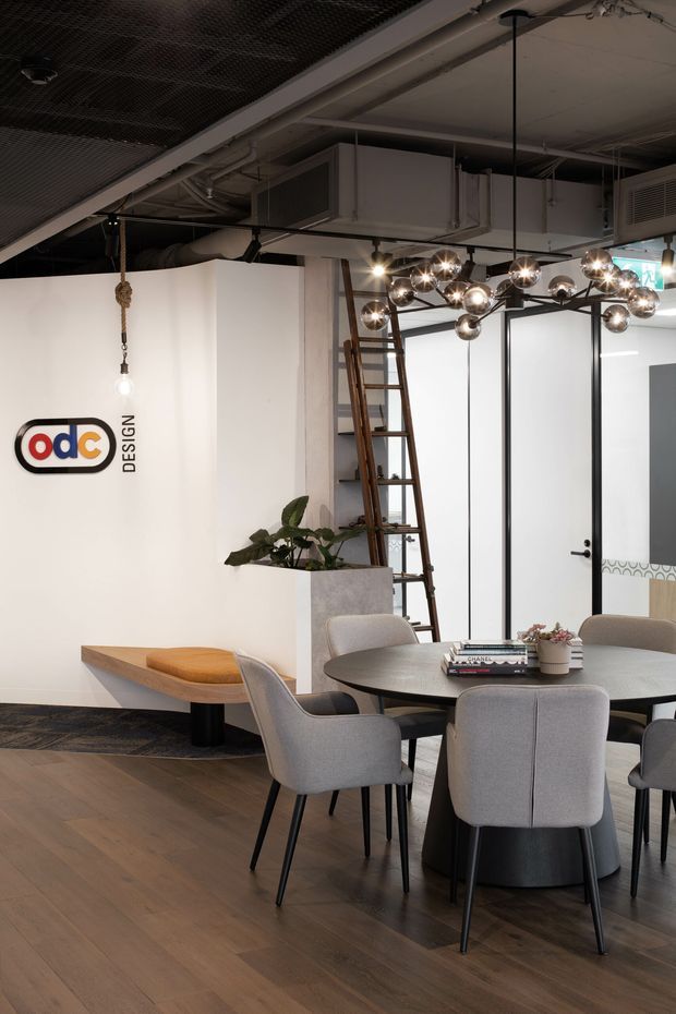 A stylish commercial office or design studio interior with an exposed industrial ceiling, visible ductwork and services, a sculptural spherical pendant light, round meeting table with upholstered chairs, timber flooring, built-in bench seating, indoor planting, and a wall-mounted design logo. A timber ladder leans against the rear wall, adding to the workshop-like character.