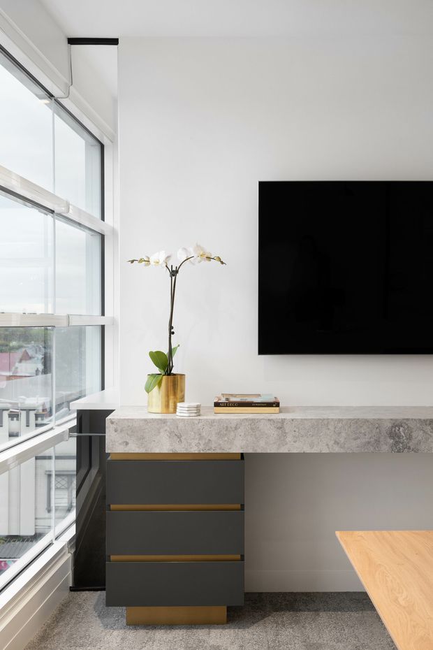 A bright contemporary living area or media space beside floor-to-ceiling glazing. A wall-mounted black television hangs above a floating grey stone console with dark built-in drawers and warm timber accents. A small orchid and books sit on the console, while a timber tabletop is visible in the foreground. The room has white walls, abundant natural light, and grey carpet flooring.