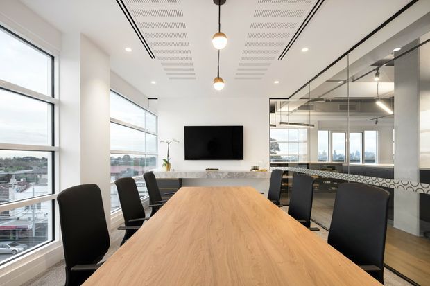 A bright contemporary office meeting room with a large light-wood conference table and black upholstered chairs. Floor-to-ceiling windows provide expansive city views, while a glass-partitioned workspace, wall-mounted screen, stone-look credenza, linear ceiling detailing, recessed lighting, and pendant lights define the fit-out.