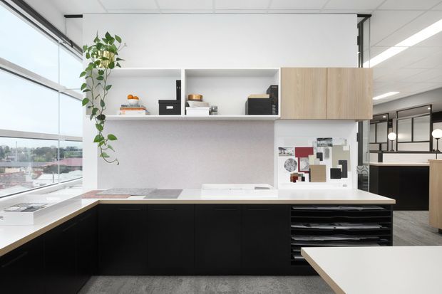 Bright contemporary office kitchenette or design studio workspace with a black L-shaped base cabinet run, pale wood overhead cabinetry, white open shelving, and a light benchtop. Large windows provide abundant daylight and views outside. A material and colour sample board is displayed on the splashback wall, with shelving, appliances, and a nearby workstation visible in the background.