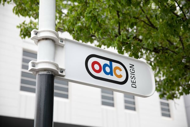 Close-up exterior view of a white directional sign mounted to a black-and-white pole, displaying the 'odc DESIGN' logo. A modern white building facade with narrow horizontal windows and leafy trees appears in the background.