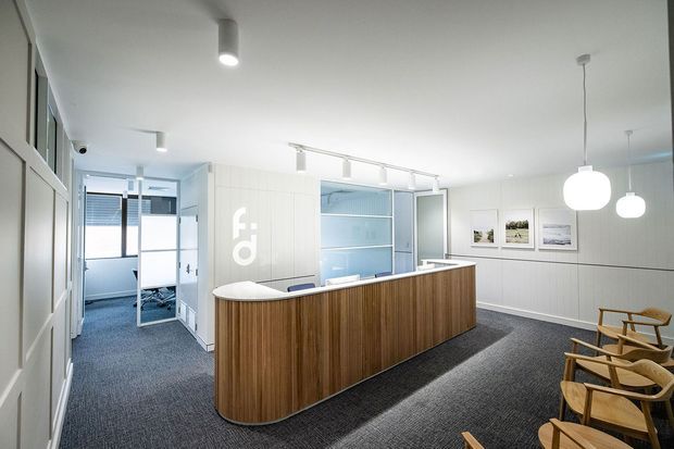 Bright contemporary reception area with a curved timber-fronted reception desk, white paneled walls, built-in display shelving, framed artwork, pendant globe lights, and a carpeted floor. Glazed partitions and an open doorway reveal adjacent office spaces, while timber lounge chairs line the right side.