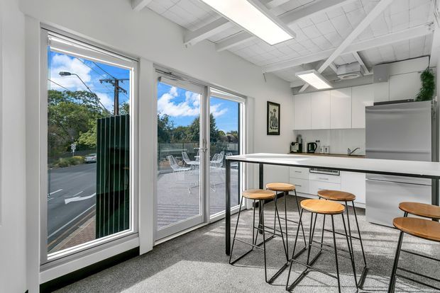 Bright contemporary kitchen and dining area with a long island bench, timber-topped black stools, white cabinetry, stainless-steel refrigerator, grey carpet, and large glazed doors opening to an outdoor deck. The space has an exposed white structural ceiling with linear fluorescent lighting and views toward a suburban street and greenery.