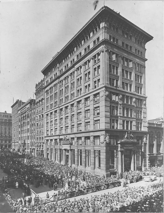 Historic black-and-white photograph of a large multi-storey commercial building on a prominent urban corner, featuring an ornate classical facade with pilasters, columns, decorative cornices, sculptural details, and a heavy projecting roofline. A very large crowd fills the surrounding streets and sidewalks.