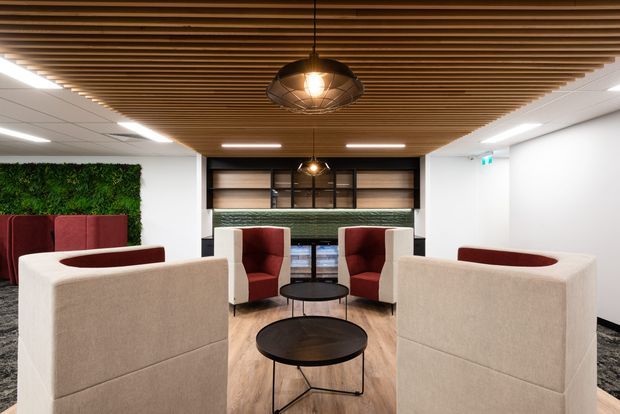 Contemporary commercial lounge and reception area with high-backed upholstered seating arranged around round black coffee tables. The space features a warm timber slatted ceiling with pendant lights, light timber-look flooring, a dark built-in shelving and service counter at the rear, white walls, and a green living wall accent.