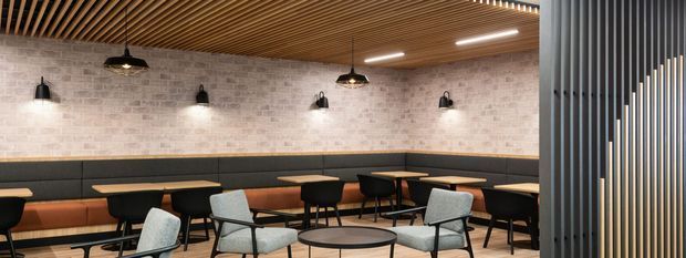 A contemporary commercial lounge or dining area with long built-in banquette seating, individual tables, upholstered chairs, exposed pale brick walls, and a timber slatted ceiling. Black pendant lights and wall sconces provide warm illumination, while a vertical dark slatted screen partially encloses the space at right.
