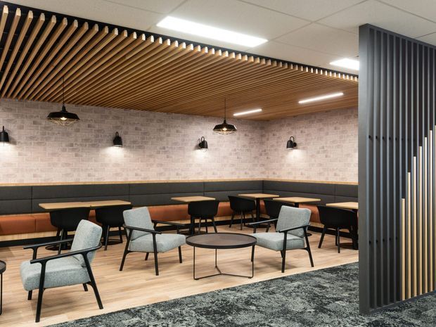 A contemporary commercial lounge or café-style breakout area with banquette seating, small dining tables, upholstered armchairs, and a round coffee table. The space features a pale exposed-brick wall, black wall sconces and pendant lights, a warm timber slatted ceiling, light timber-look flooring, and a dark vertical slatted divider at the entrance. A patterned dark rug defines the seating zone.