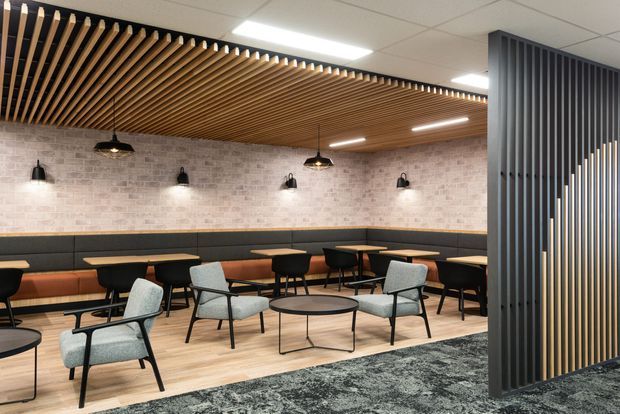 Contemporary commercial lounge or breakout dining area with upholstered booth seating, café tables, lounge chairs and pendant lighting. The space features a light timber floor, pale exposed-brick-look wall finish, a slatted timber ceiling feature, and a dark vertical slatted screen creating separation at the right.