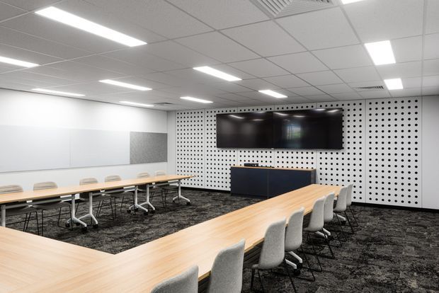Modern commercial meeting or training room with long light-wood conference tables arranged in a U-shape, grey upholstered chairs, dark patterned carpet, white acoustic wall panels, a large black display screen, and a suspended grid ceiling with recessed linear lighting.