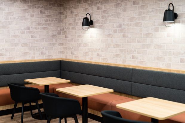 A contemporary commercial dining or hospitality interior with built-in dark upholstered banquette seating wrapping around a corner, light timber tables, black chairs, pale exposed brick walls, and black wall-mounted industrial-style sconces. The space has a warm, understated café or restaurant atmosphere.