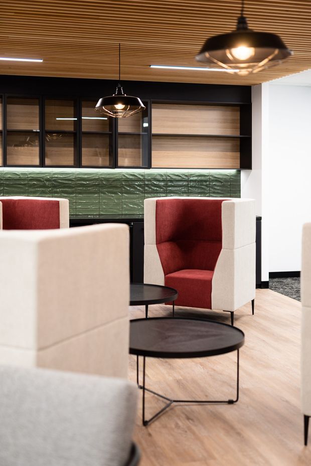 A stylish commercial lounge or reception area with cream and burgundy upholstered armchairs, black round coffee tables, pale timber flooring, dark built-in cabinetry, and a green textured tiled backsplash. The space features a slatted timber ceiling and suspended black pendant lights, creating a warm, refined contemporary interior.