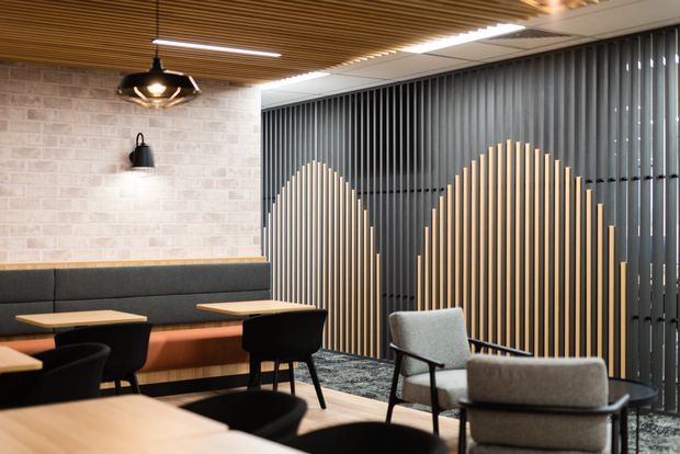 Contemporary commercial-style lounge or dining area with built-in dark upholstered banquette seating, small tables, armchairs, and a dramatic vertical slatted feature wall. The space combines a warm timber-slat ceiling, pale brick-look wall finish, black metal lighting, and a sculptural timber-and-dark-slat partition, creating a refined hospitality or office breakout atmosphere.