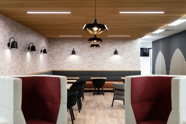 A contemporary commercial lounge or hospitality interior with booth seating, high-backed upholstered chairs, banquettes, and tables arranged beneath a warm timber slatted ceiling. The space features pale brick-look wall finishes, black wall sconces and pendant lights, light timber-look flooring, and a dark vertical feature screen with arched detailing.
