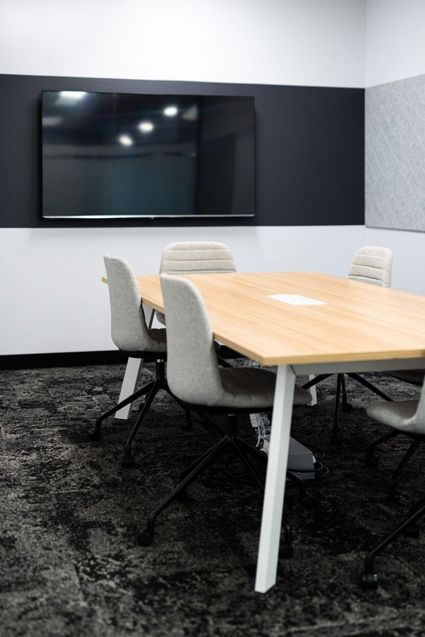 A contemporary meeting room with a large light-wood conference table surrounded by upholstered swivel chairs. A wall-mounted flat-screen television is set against a dark charcoal feature wall, with a grey acoustic-textured panel on the adjacent wall. The room has white walls, black skirting, and dark patterned carpet.