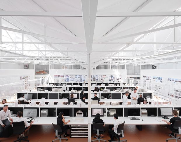 Bright, expansive open-plan commercial workspace with rows of white workstations, shelving, display boards, computers, and employees working at desks. The space has a high white exposed structural roof with extensive trusses and beams, abundant daylight, and dark timber flooring. The image resembles a design studio, showroom, or architectural office.