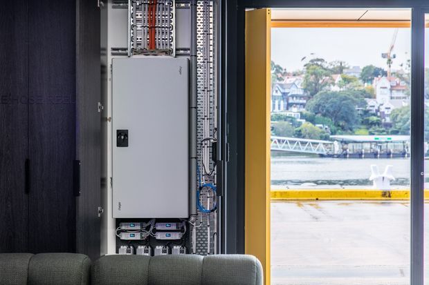 Interior view of a contemporary commercial or industrial space showing an exposed electrical services panel and neatly organized wiring beside a large glazed opening. A dark built-in wall and the top of a sofa are visible in the foreground, while an urban waterfront or dockside setting can be seen outside.