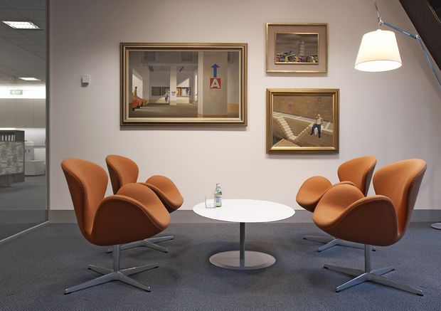 A refined lounge or waiting area with four sculptural rust-orange leather armchairs arranged around a round white pedestal table. The space has grey carpet, pale grey walls, framed artwork, a suspended pendant light, and a glazed opening to an adjacent office or corridor. The overall setting appears polished and contemporary, with a calm, professional atmosphere.
