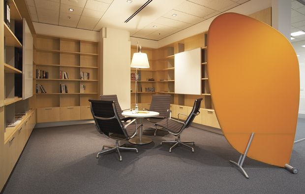 Contemporary office meeting area with a round table and leather swivel chairs, surrounded by extensive built-in light timber shelving and cabinetry. A large curved orange freestanding acoustic privacy screen dominates the right side, while grey carpet and a suspended ceiling create a practical commercial interior.