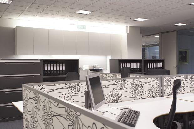 Modern open-plan office interior with white workstations, patterned privacy screens, computer monitors, filing cabinets, shelving with binders, and a suspended acoustic-tile ceiling. The space is brightly lit and arranged as a compact commercial workspace.