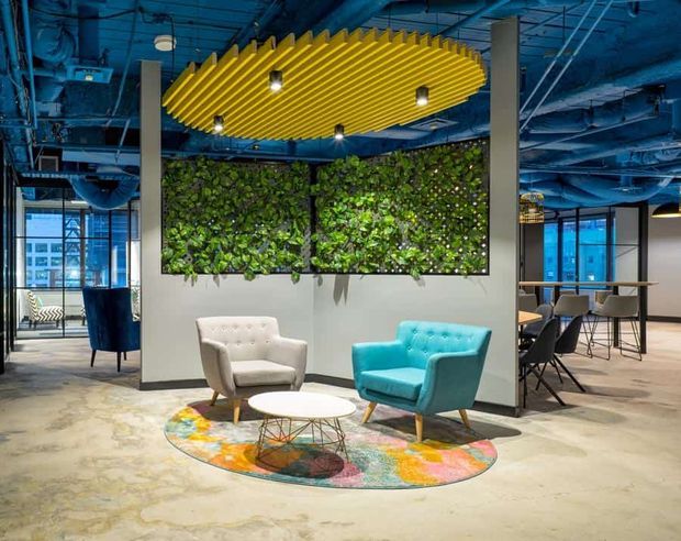 A vibrant contemporary commercial lounge or reception area with two tufted armchairs, a round coffee table, colourful patterned rug, and a large living plant wall. The space features exposed blue-painted services and structure, glazed meeting or dining areas, concrete-look flooring, and a distinctive suspended yellow slatted ceiling feature with integrated lighting.