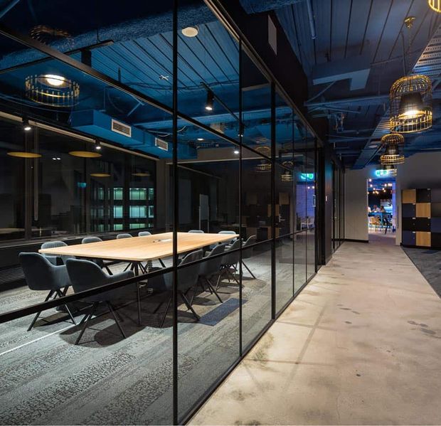 A dark, contemporary commercial office interior featuring a long meeting room enclosed by black-framed floor-to-ceiling glass partitions. The room contains a large light timber conference table with dark upholstered chairs, patterned carpet, exposed blue-painted ceiling services, suspended track lighting, and distinctive cage-like pendant lights. A polished concrete corridor runs alongside the glazed meeting room, with a reception or breakout area visible in the distance.