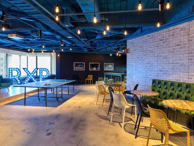 A lively industrial-style commercial interior with exposed blue-painted ceiling services, pendant filament lights, polished concrete flooring, a white brick feature wall, and casual dining tables along a patterned upholstered banquette. A table-tennis table and illuminated sign occupy the open activity area.