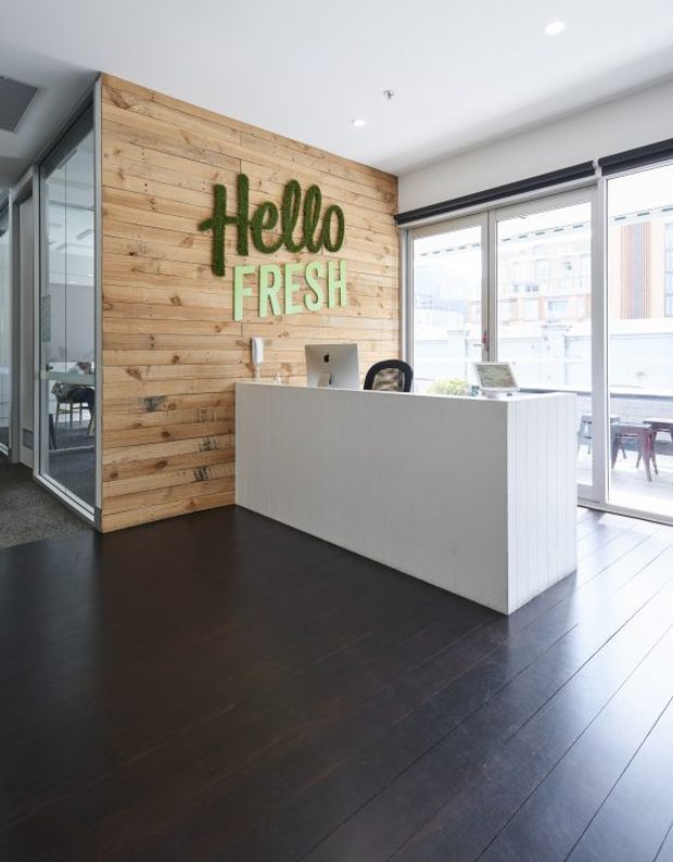 A bright contemporary commercial reception area with a white reception desk, computer workstation, dark timber flooring, and a full-height light timber feature wall bearing a green “Hello Fresh” sign. Floor-to-ceiling glazing opens the space to a bright exterior, while a glazed meeting room is visible to the side.