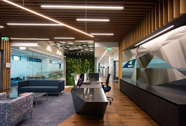 Contemporary commercial office interior with an open reception or collaborative workspace. The space features a long angular dark workstation, black cabinetry, upholstered lounge seating, glass-partitioned meeting rooms, timber flooring, a dramatic faceted geometric wall installation, and a timber-slat ceiling with linear integrated lighting. A living green wall adds a natural accent.