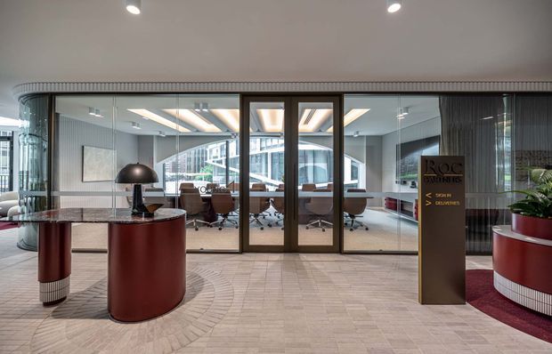A sophisticated commercial office reception and entry area viewed from the lobby. Curved glazed meeting-room walls and framed glass doors reveal a contemporary boardroom with a long table and rolling office chairs. The lobby features sculptural rounded reception furniture in burgundy and dark timber tones, a freestanding branded directory, patterned carpet flooring, curved ribbed wall detailing, recessed linear ceiling lighting, and a planted pot.
