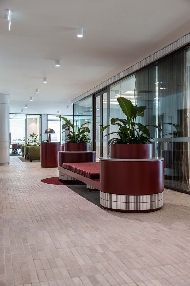 A bright contemporary commercial interior lobby or communal office space with a long corridor, floor-to-ceiling glazed meeting-room partitions, pale rectangular tiled flooring, and a white ceiling with recessed downlights. Large burgundy-red cylindrical planter and seating units with ribbed light-coloured bases form the main feature, containing tall green plants. Additional lounge seating and glazed rooms extend into the background.