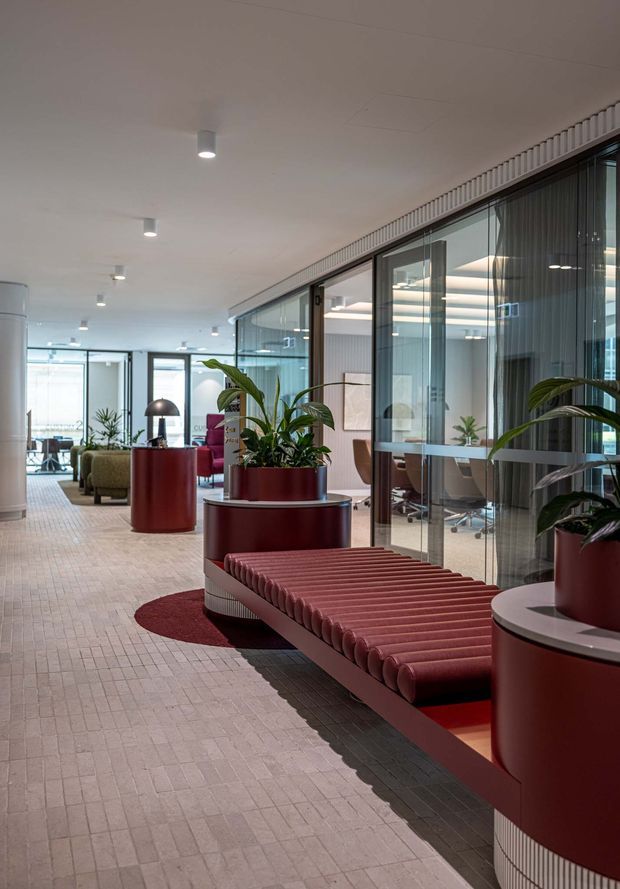 A contemporary commercial office interior with a long circulation lounge area. The space features glass-partitioned meeting rooms, pale tiled flooring, a white ceiling with recessed downlights, burgundy cylindrical planters and tables, abundant indoor greenery, and a ribbed burgundy upholstered bench. Office workstations and chairs are visible behind the glazing.